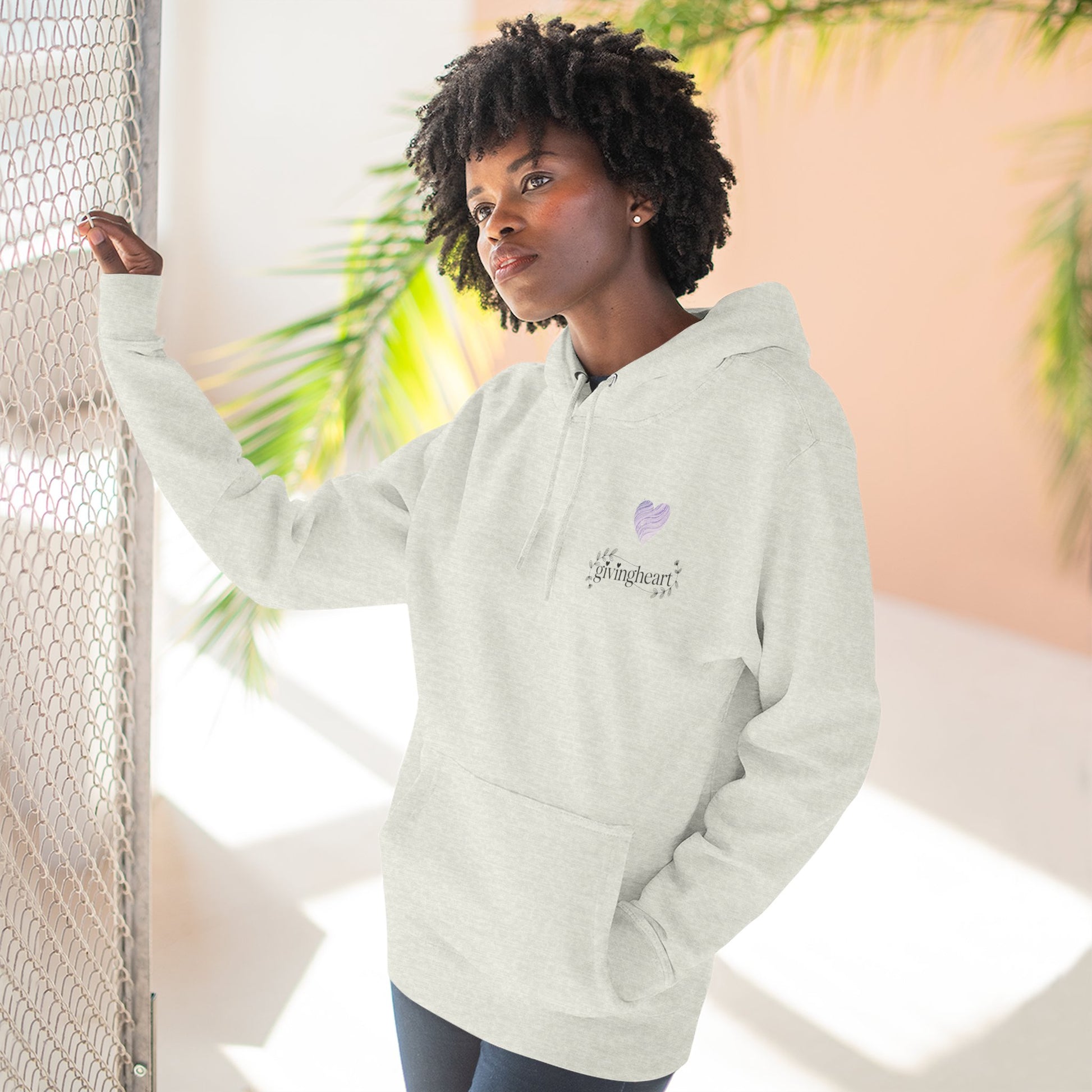 Person wearing a light gray hoodie with a lilac colored heart logo standing against a neutral background with plants.