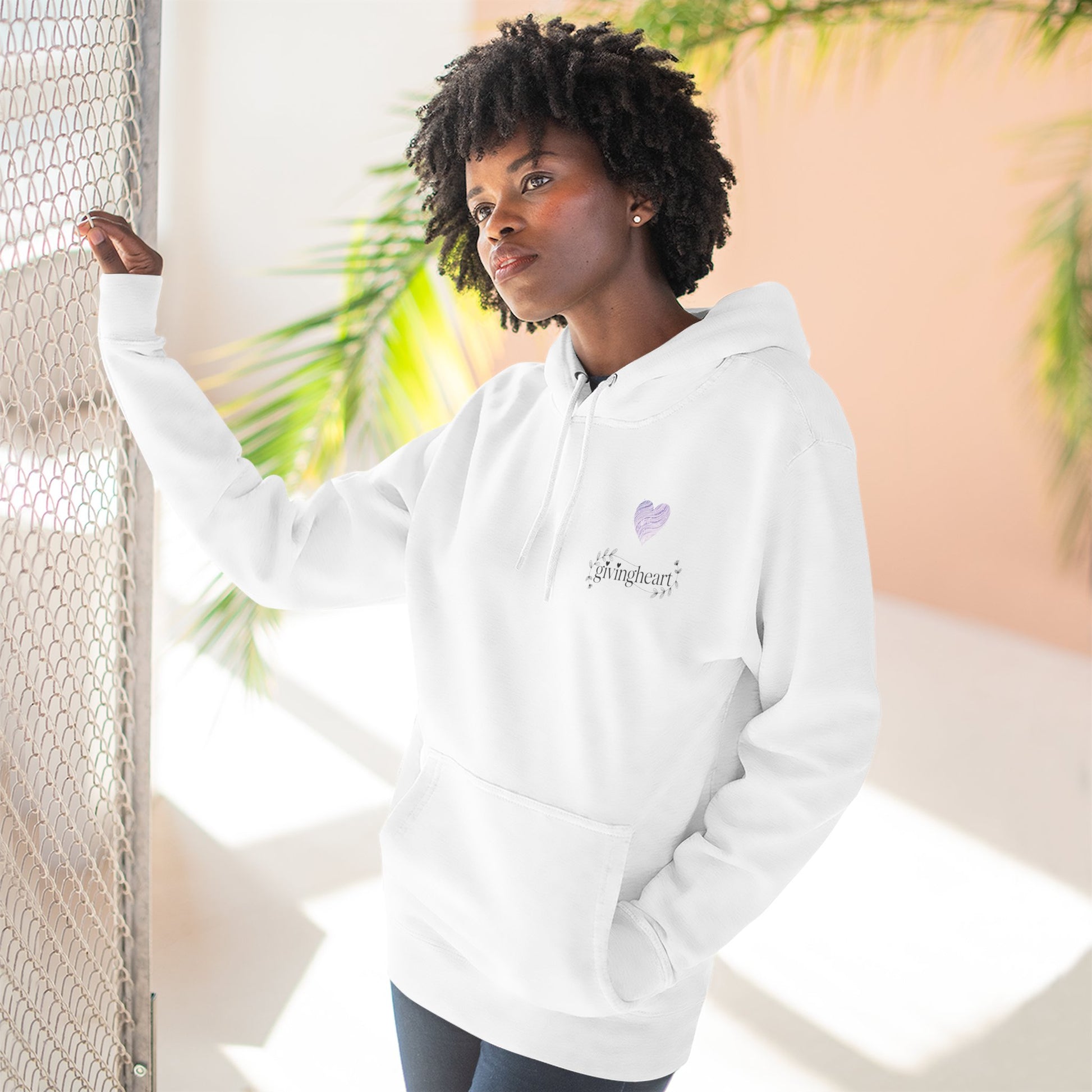 Person wearing a white hoodie with a lilac colored heart logo, standing against a light background with plants.