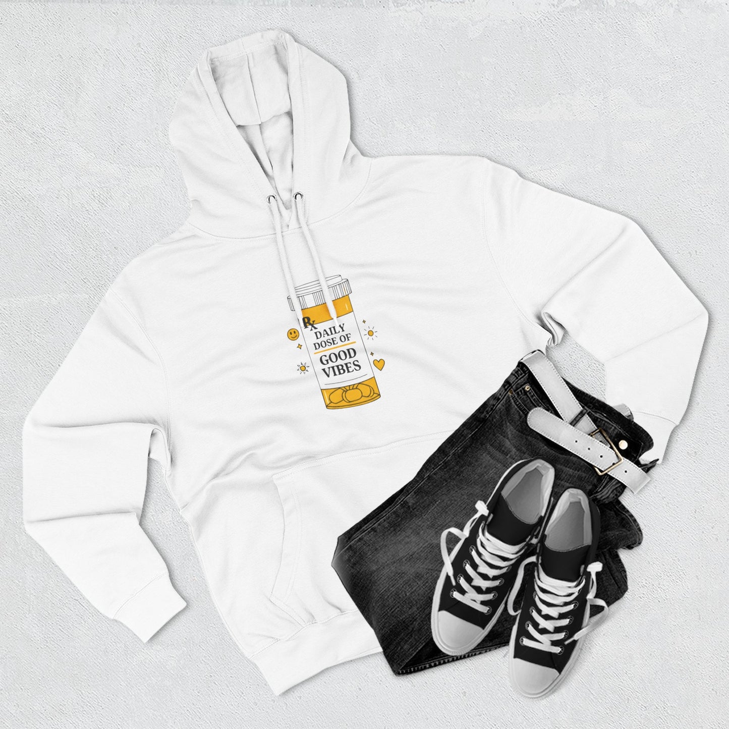 White hoodie with a daily dose of good vibes design, black jeans, and black sneakers on a light gray background